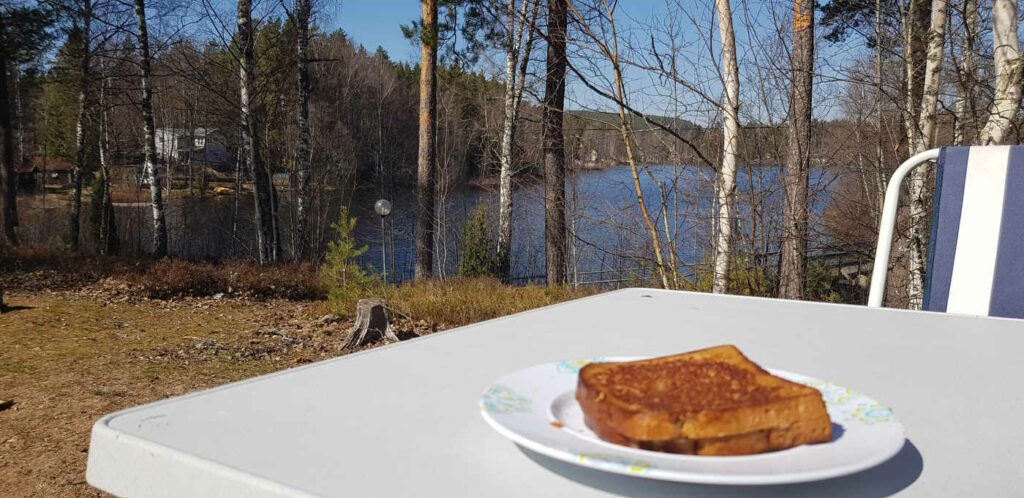 Unscharf im Vordergrund: Ein Käsetoast liegt auf einem Teller auf einem Campingtisch. Dahinter ist ein See zu sehen. Die Sonne scheint.